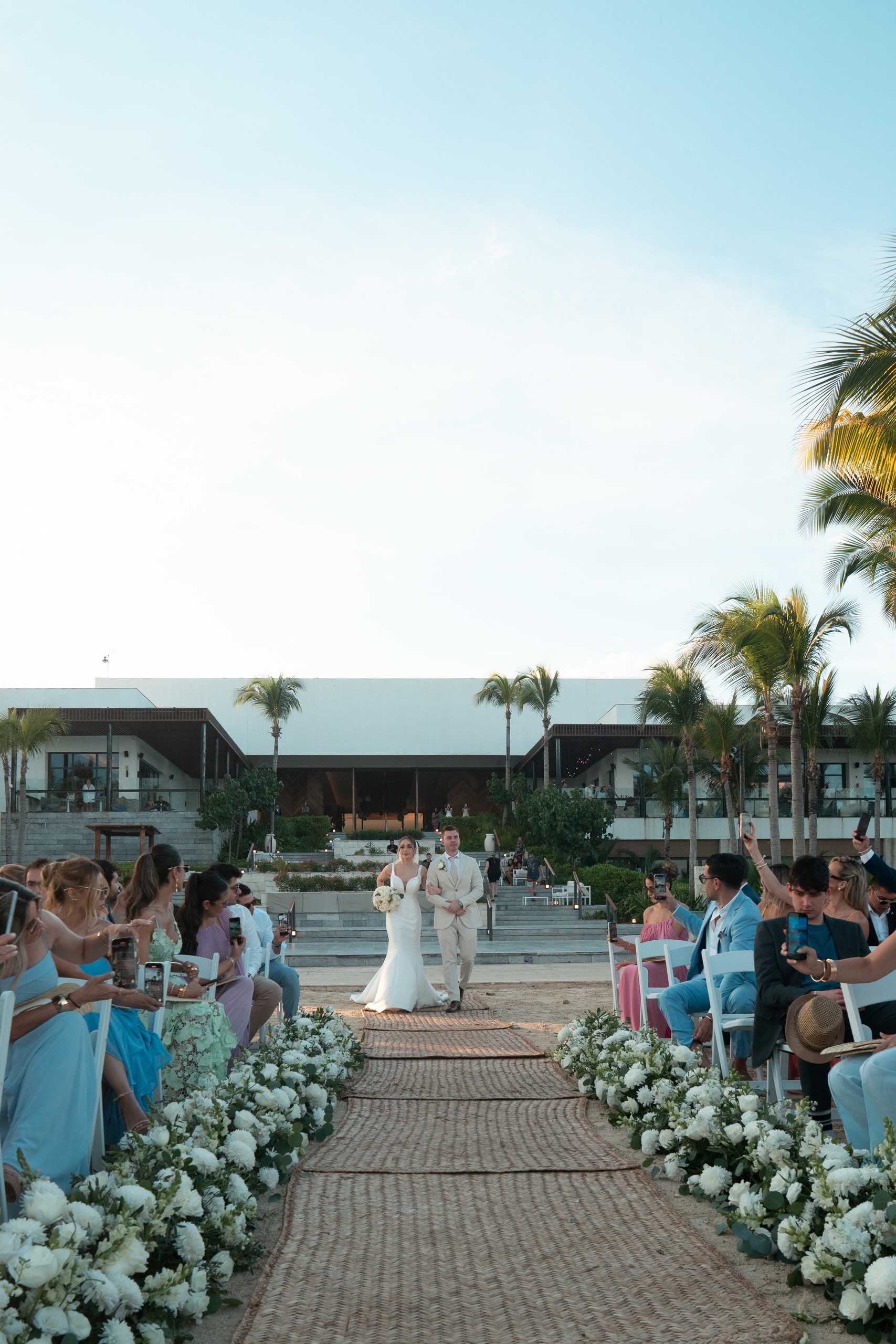 Los novios entrando por el pasillo de la ceremonia entre palmeras y flores blancas, frente al mar